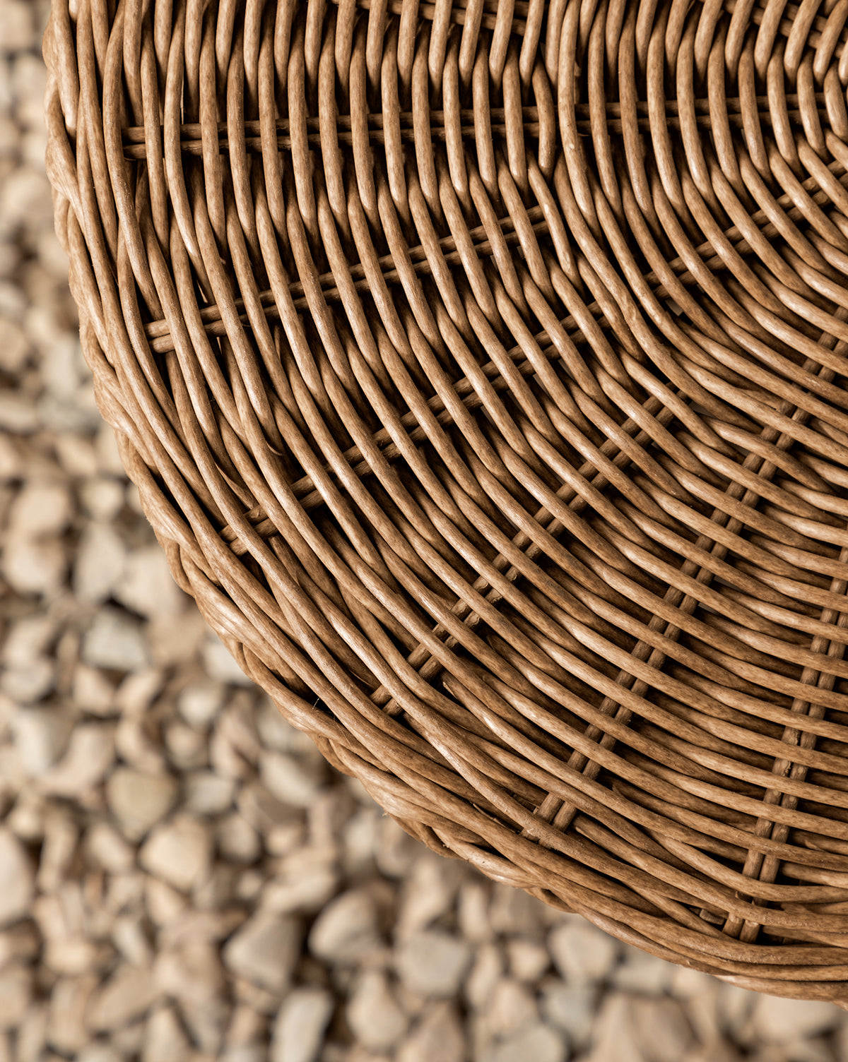 Close-up of the round Haviland Side Table in woven wicker on the upper right, with its textured pattern sharply detailed. A blurred background of light pebbles enhances this perfect piece for outdoor lounging, by Vendixs