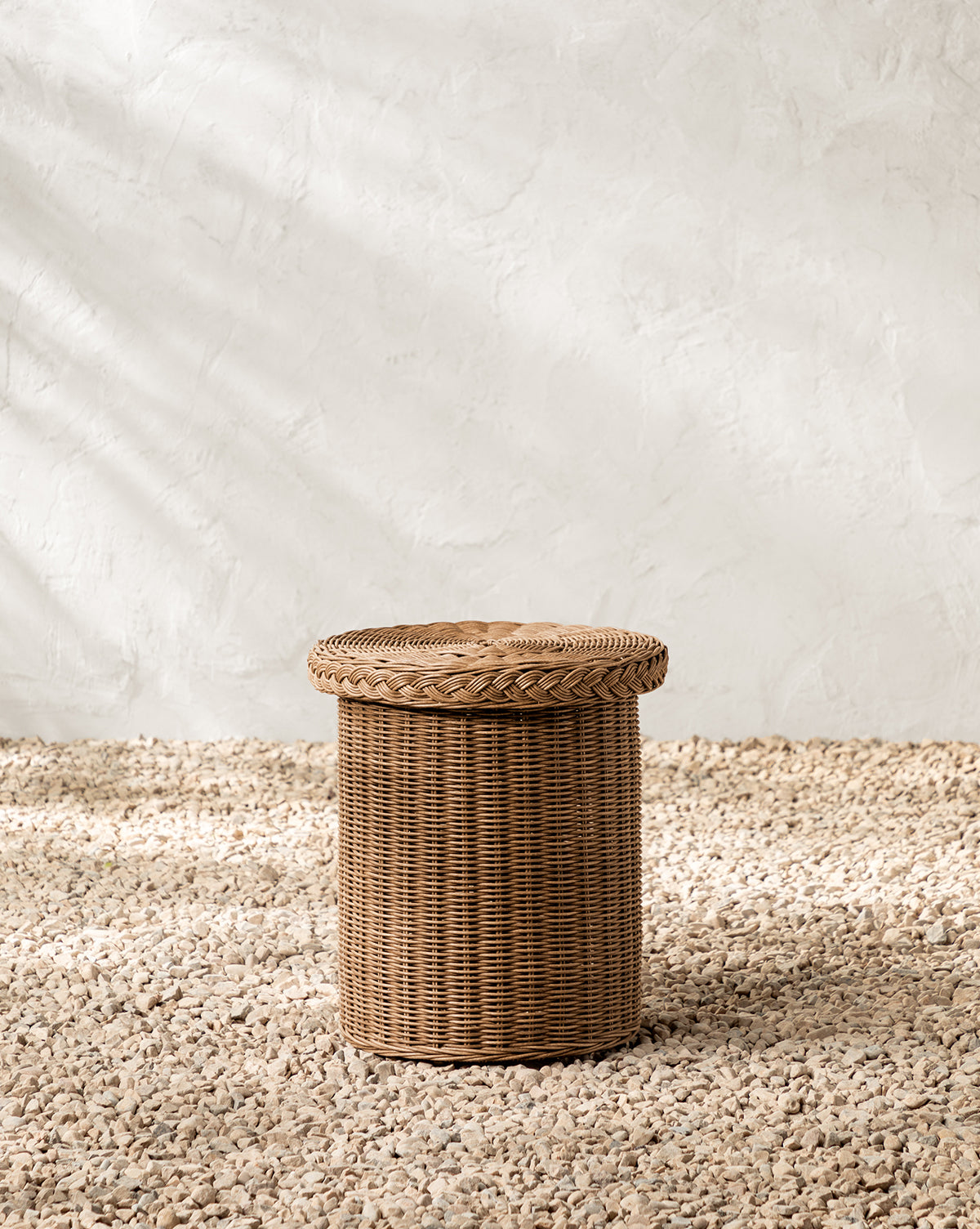 The Haviland Side Table, a round wicker basket with a lid, stands on gravel by a textured white wall. Its weather-resistant design and warm sunlight make it perfect for outdoor lounging and versatile as both storage and side table, by McGee & Co.