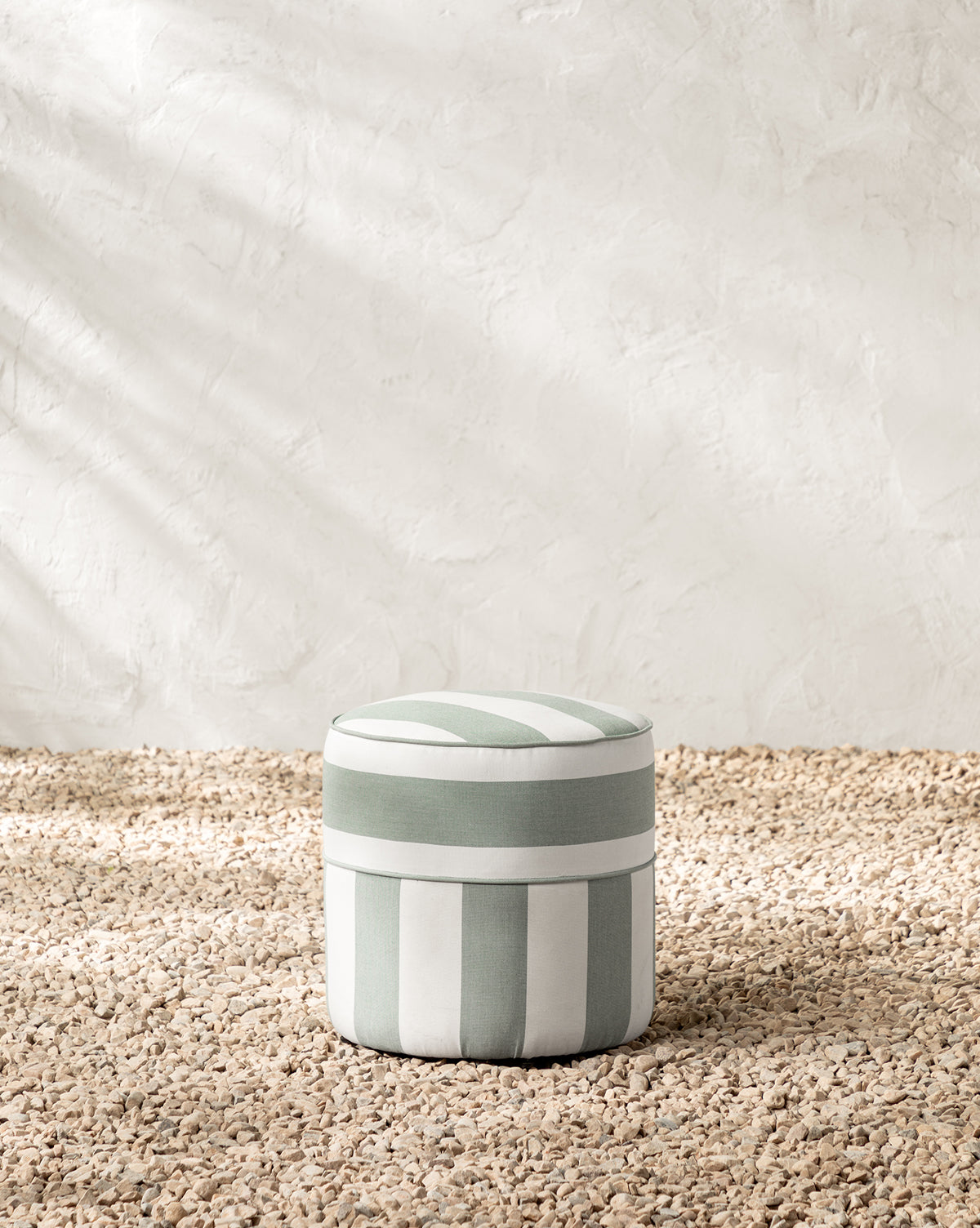 The Makerspalm Haviland Striped Ottoman features light green and white vertical stripes, sits on a pebble floor by a textured white wall, and is weather-resistant—perfect as a stylish outdoor accent. Vendixs