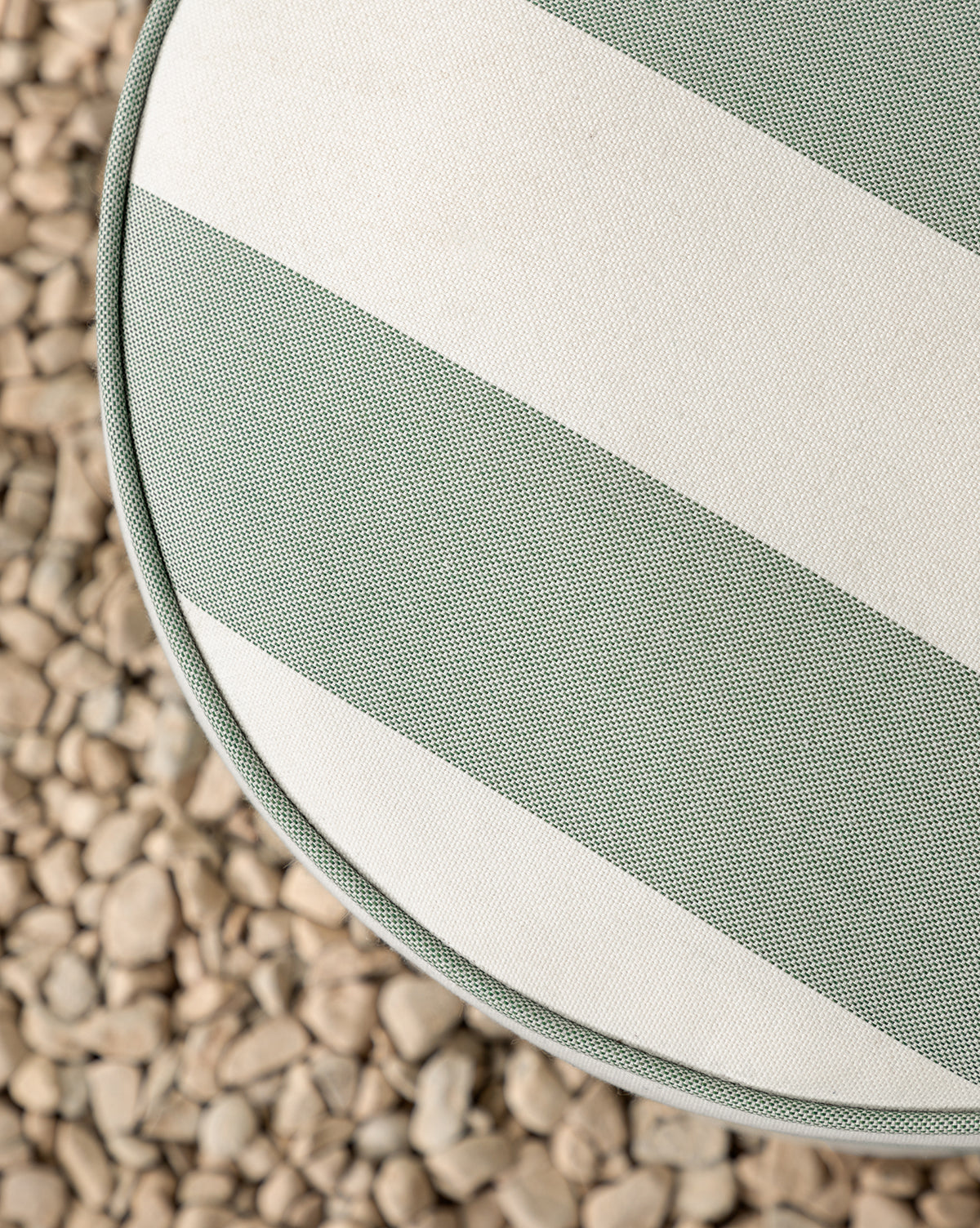 The Makerspalm Haviland Striped Ottoman, featuring wide green and white weather-resistant stripes, is shown from above on a bed of small, light-colored pebbles, with a partial view of its cushion and the textured surface. Vendixs
