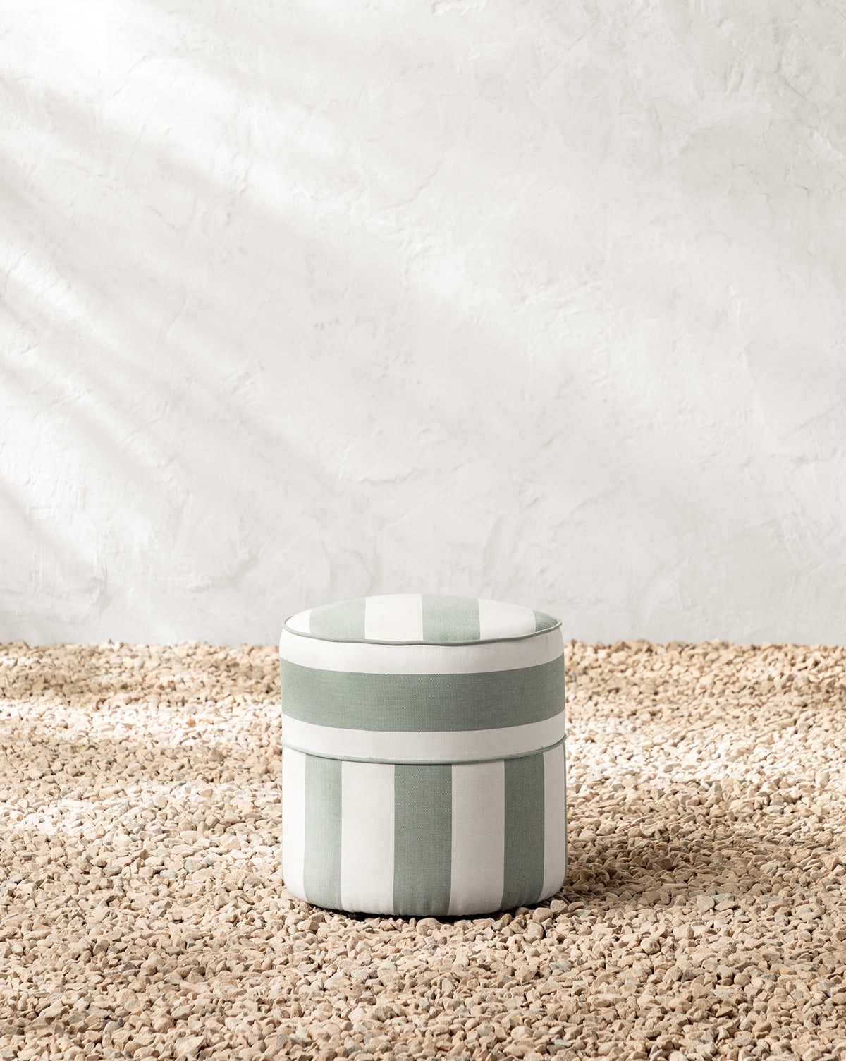 The Makerspalm Haviland Striped Ottoman in light green and white sits on a textured pebble floor by a white wall, with sunlight casting soft shadows that highlight this weather-resistant outdoor ottoman’s inviting style. Vendixs