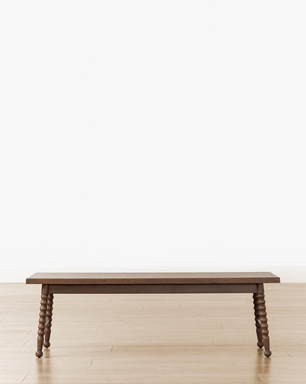 A Heathcliff Bench with turned legs sits on a light wood floor against a plain white wall, by Vendixs