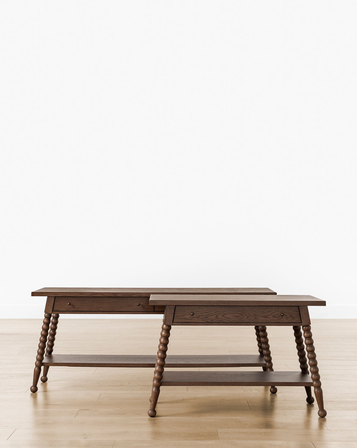 Two Heathcliff Console tables of varying heights feature turned bobbin legs, a lower shelf, and a single drawer each. Their subtle walnut oak finish highlights classic charm against a plain white background and light wood floor, by Vendixs