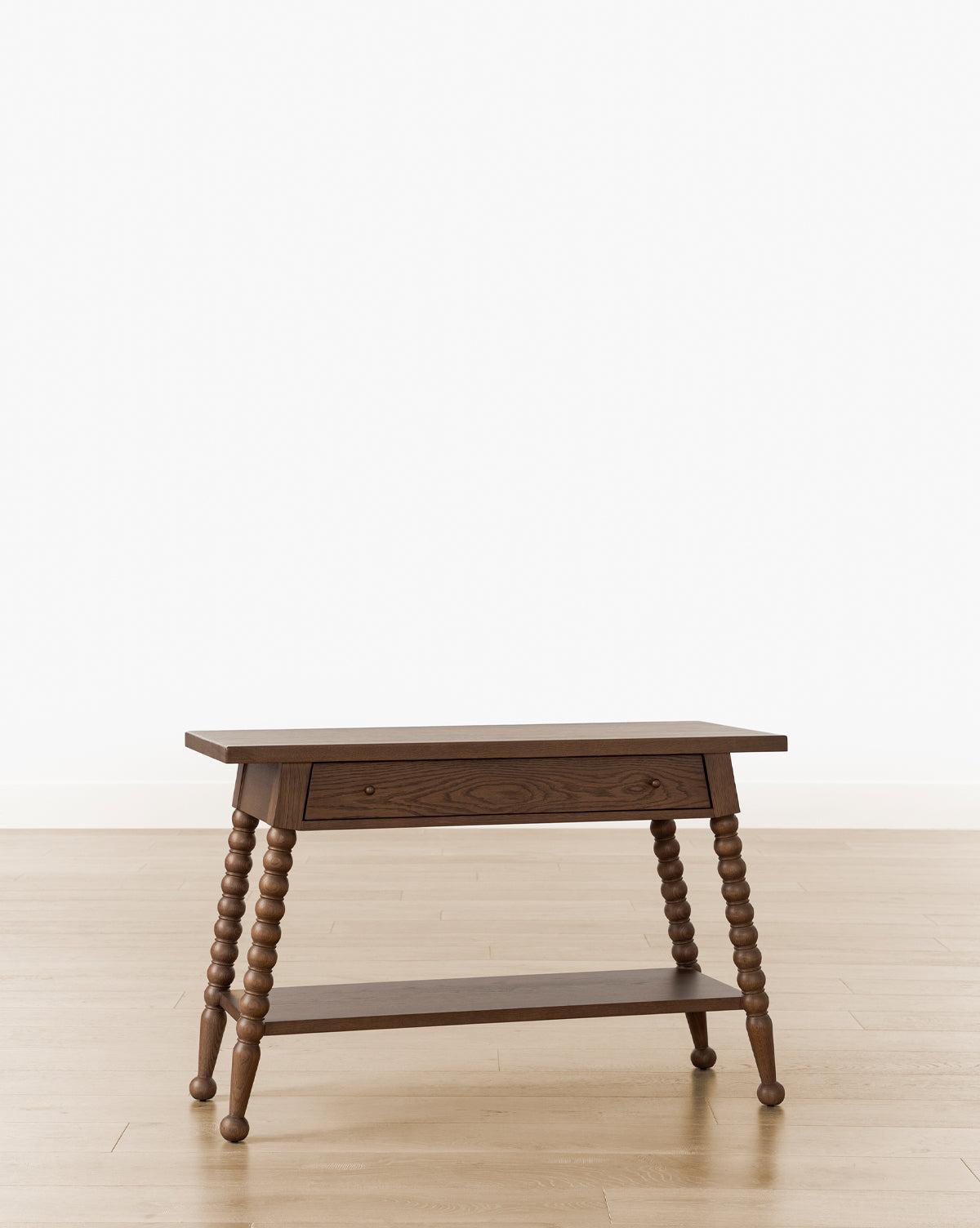 The Heathcliff Console is a wooden side table featuring turned bobbin-style legs, a single front drawer, and a lower shelf, shown on a light wood floor against a plain white background, by Vendixs