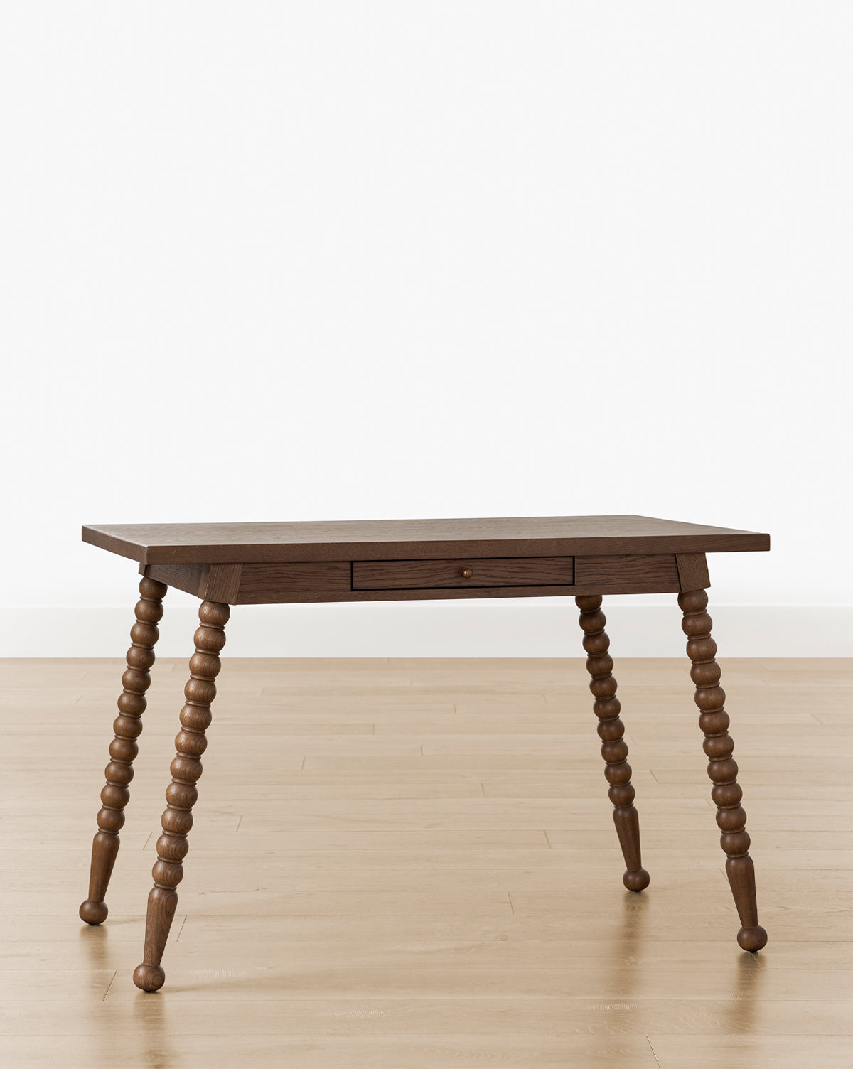 The Heathcliff Desk features a bobbin-style design with a central drawer and four spindle-turned legs, shown on a light wood floor against a plain white background by Vendixs