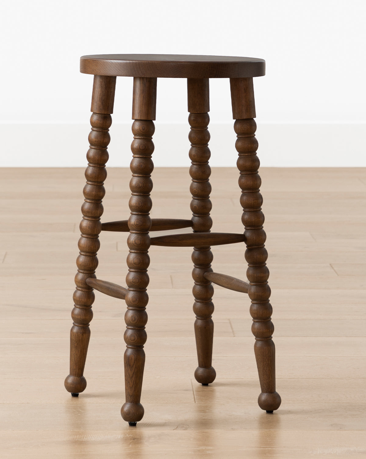 A Makerspalm Heathcliff Stool with a round wooden seat and four spindle-style legs in walnut oak finish, sits on a light wood floor against a white wall, Vendixs