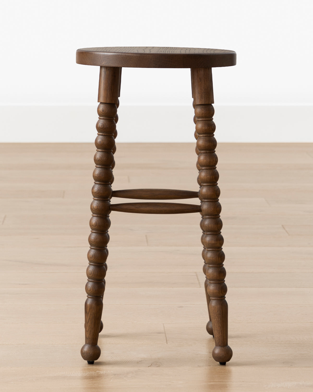 The Makerspalm Heathcliff Stool, featuring a walnut oak finish, turned spindle legs, and a round seat, is placed on a light wood floor against a white wall. Vendixs