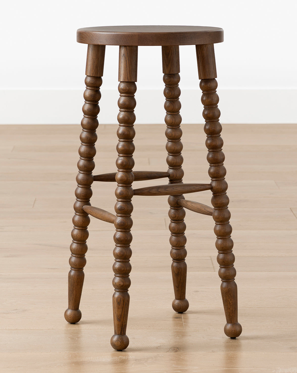 The Makerspalm Heathcliff Stool features a walnut oak finish, four turned legs with crossbars, and a round wooden seat, shown on a light wood floor against a plain white wall. Vendixs