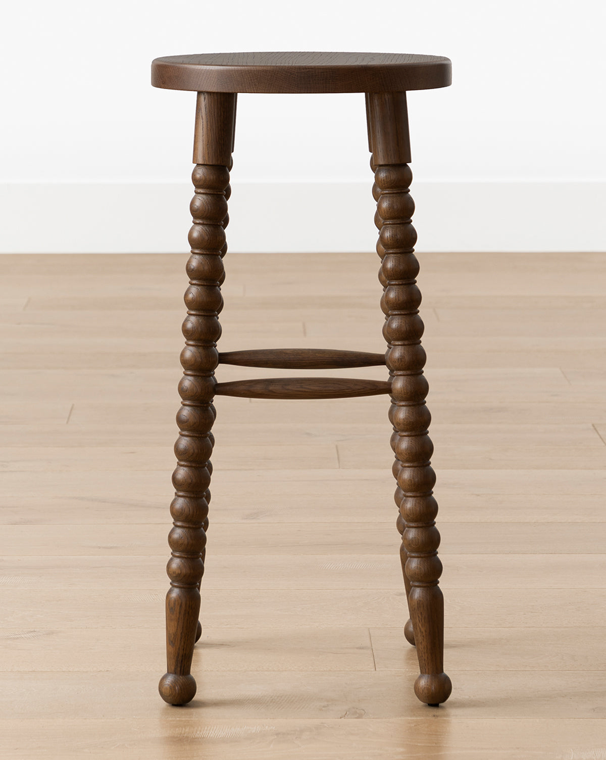 The Makerspalm Heathcliff Stool features a dark walnut oak finish, three intricately turned legs, and a round seat, shown on a light wood floor against a white wall. Vendixs