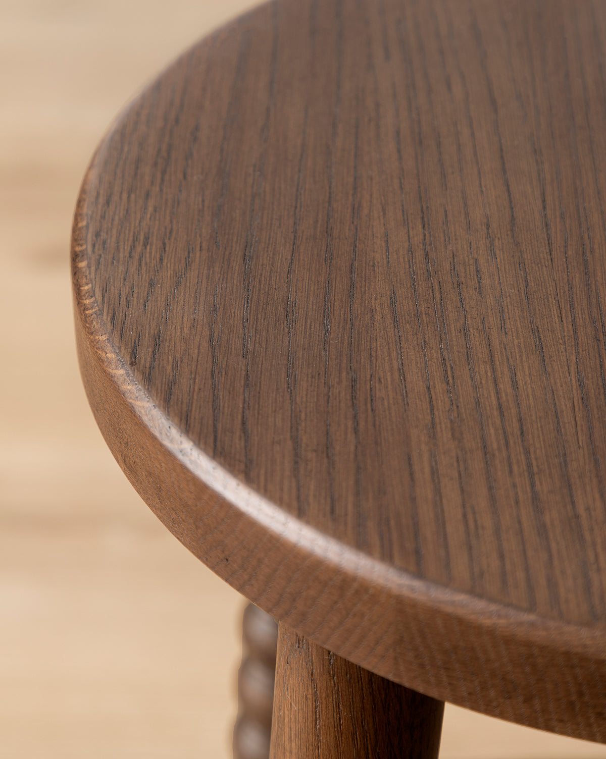 Close-up of a Makerspalm Heathcliff Stool shows its round tabletop with a smooth, dark brown walnut oak finish and visible wood grain, plus one distinctive bobbin-style leg—finished in the refined style of Vendixs