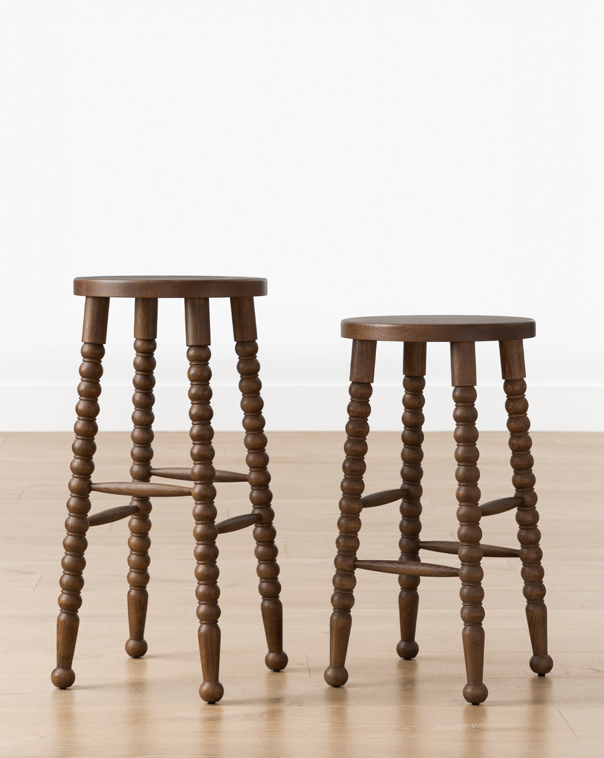 Two Makerspalm Heathcliff Stools with round seats and bobbin-style legs in walnut oak finish are set on a light wood floor against a plain white wall, Vendixs