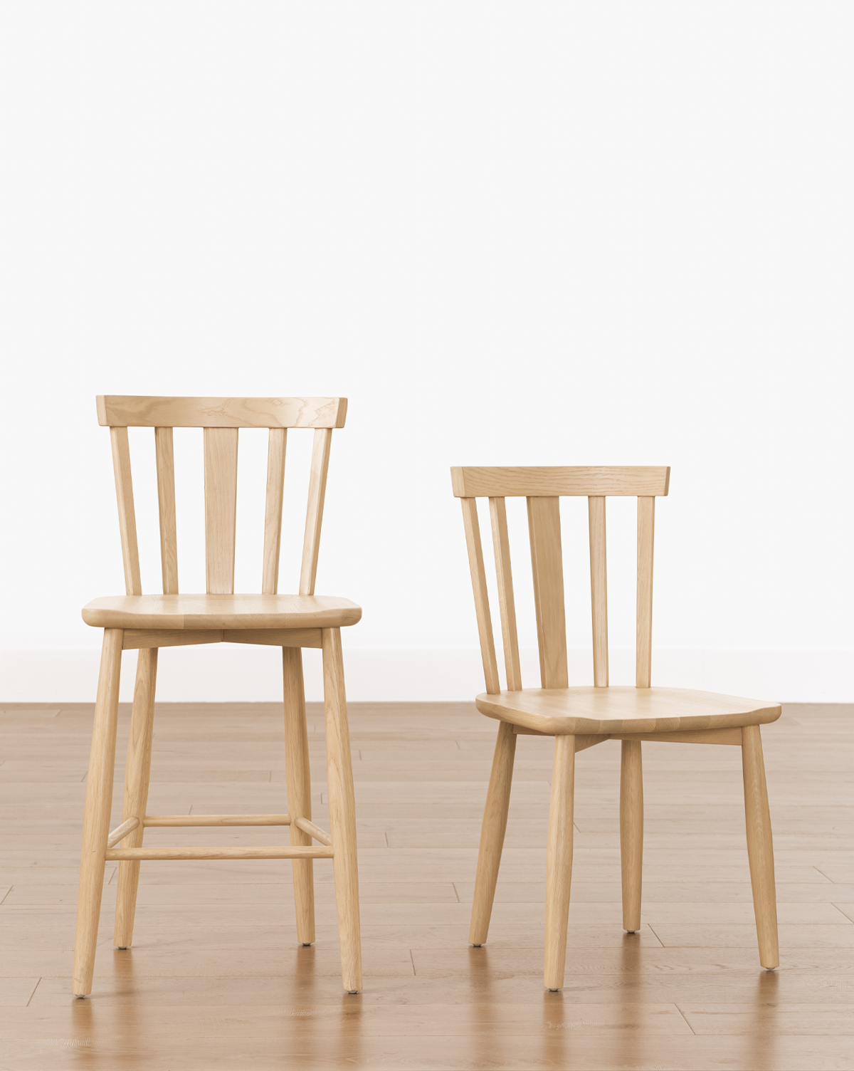 Two solid wood chairs, including a tall Hemingway Stool and a shorter chair, both with vertical slatted backrests, are side by side on a light wood floor against a plain white background, by Vendixs