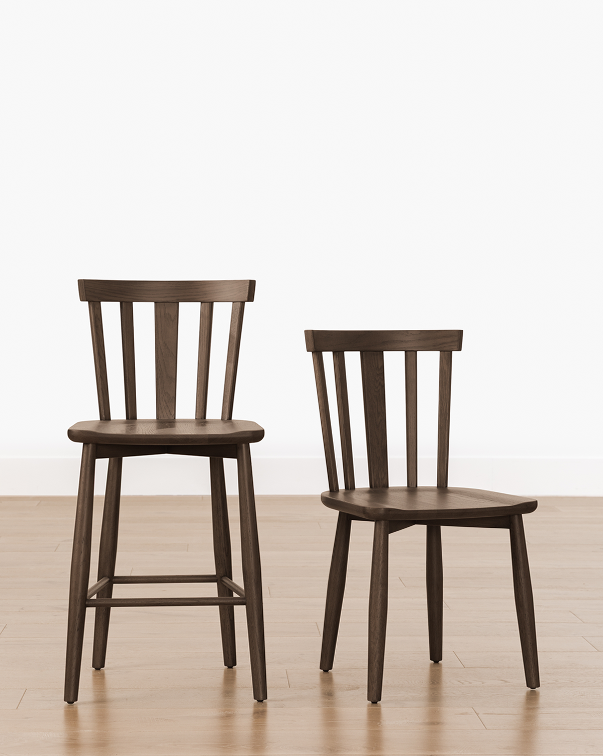 Two dark wooden chairs—a tall bar stool and the Hemingway Chair—sit side by side on a light wood floor. Featuring vertical slats and solid wood construction, they’re set against a plain white wall for a timeless look, by Vendixs
