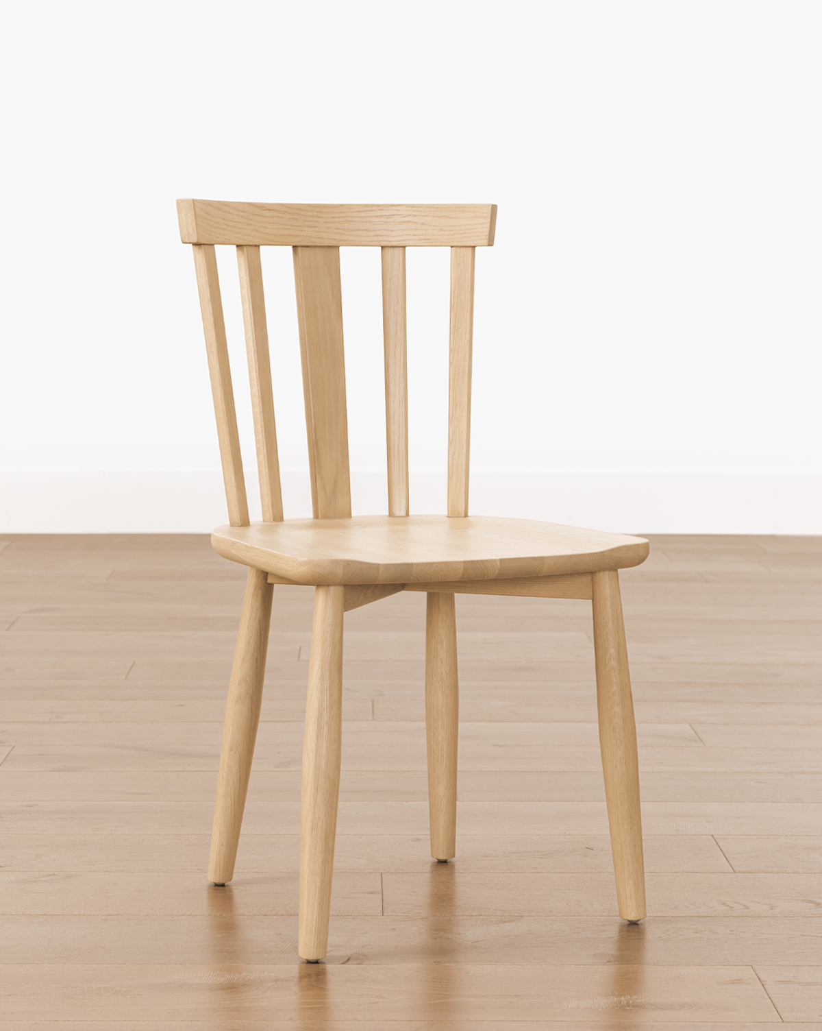 The Hemingway Chair, featuring a slatted backrest and four legs, is crafted from solid wood and stands on a wooden floor against a plain white wall, by Vendixs