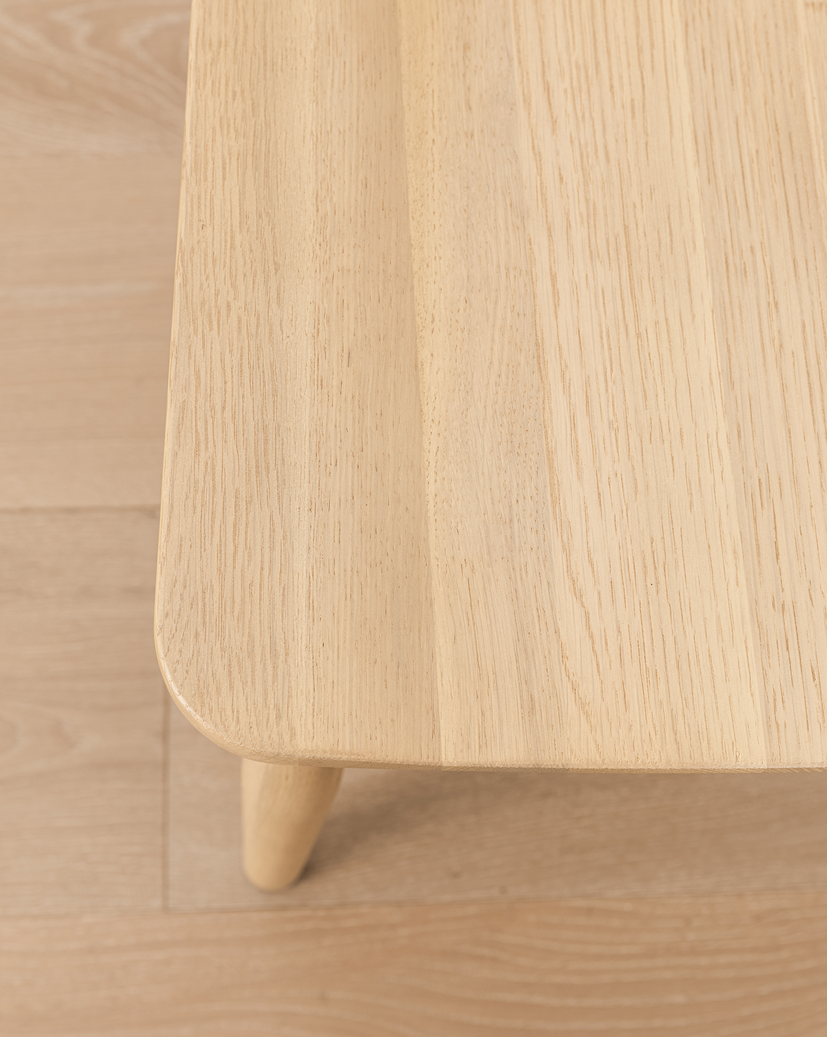 Close-up of a light wooden table with rounded corners and visible grain, set on a matching floor next to a versatile Hemingway Chair, by Vendixs