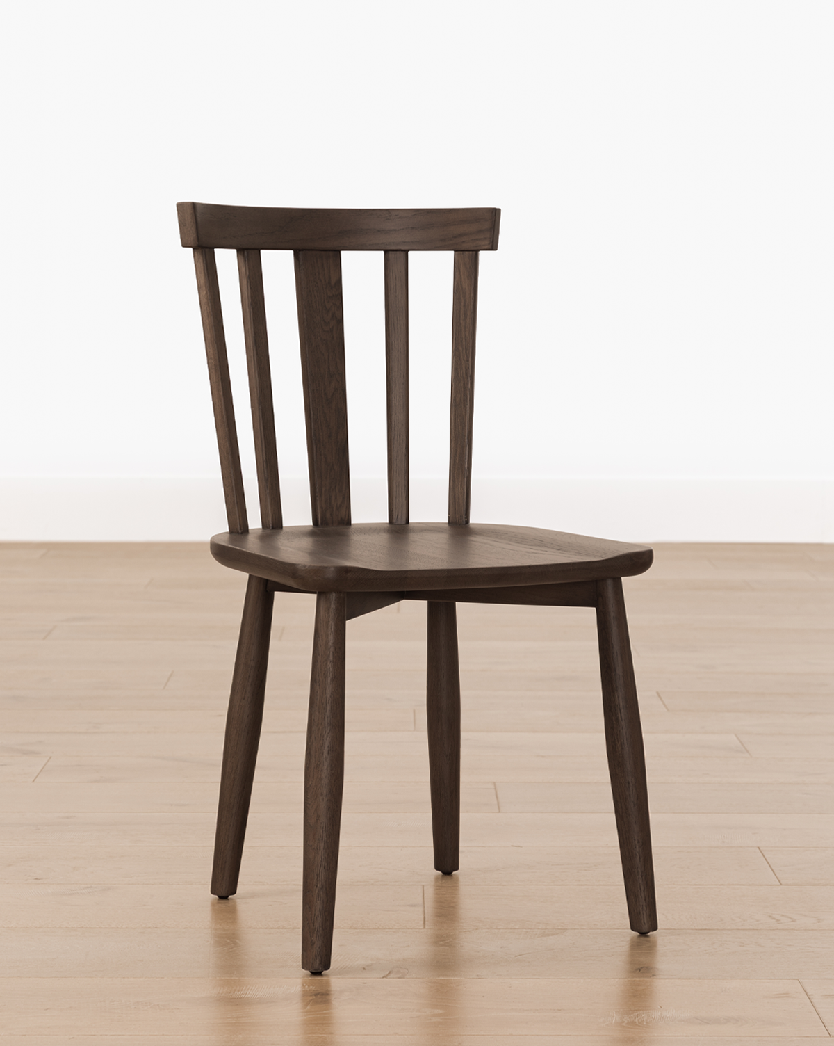 The Hemingway Chair is a versatile dining chair with a dark wooden finish, featuring a curved backrest with vertical slats. It's shown on a light wood floor against a plain white wall, by Vendixs