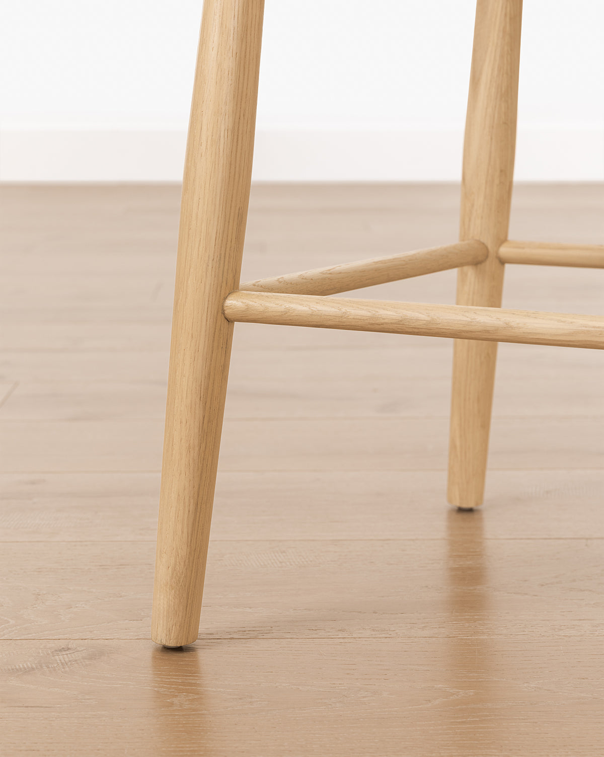 Close-up of the Hemingway Stool’s solid wood legs on a light brown wooden floor, with white baseboard and wall in the background, by Vendixs