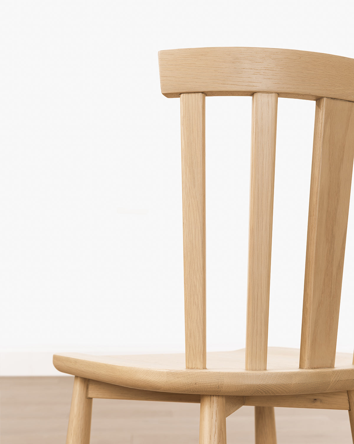 A close-up of the Hemingway Stool, crafted from solid wood with vertical back slats, stands against a plain light wall and wooden floor; only part of the stool is visible, hinting at its placement near kitchen counters, by Vendixs