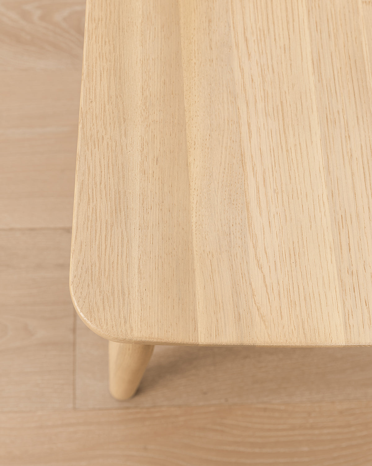 A close-up of a solid wood table with rounded edges and rich grain on a matching floor—pairs seamlessly with the elegant Hemingway Stool for any kitchen or dining space, by Vendixs