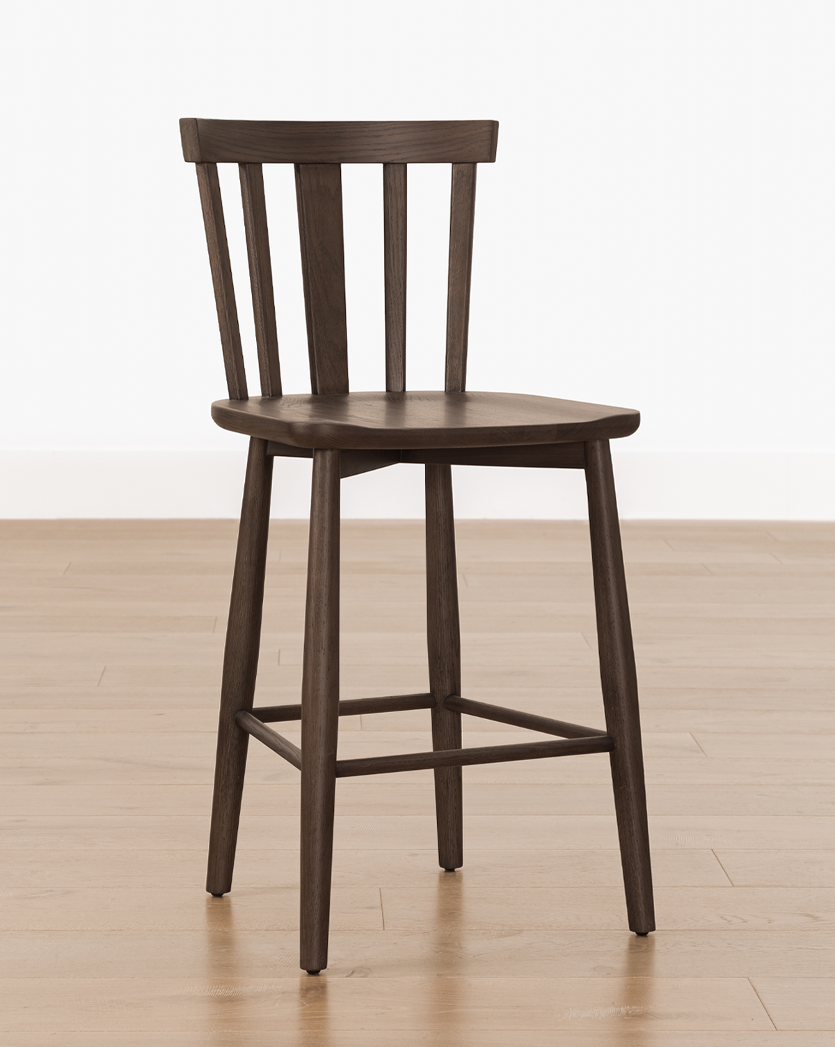 The Hemingway Stool, a dark solid wood stool with a flat seat, slightly angled legs, and a backrest featuring three vertical slats, sits on a light wood floor against a plain white wall—ideal for pairing with kitchen counters, by Vendixs
