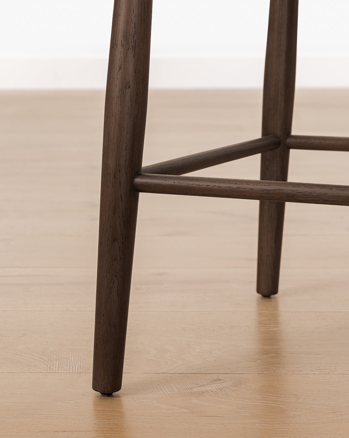 Close-up of the Hemingway Stool’s solid wood legs and crossbars on a light wood floor, ideal for kitchen counters with a white wall backdrop, by Vendixs
