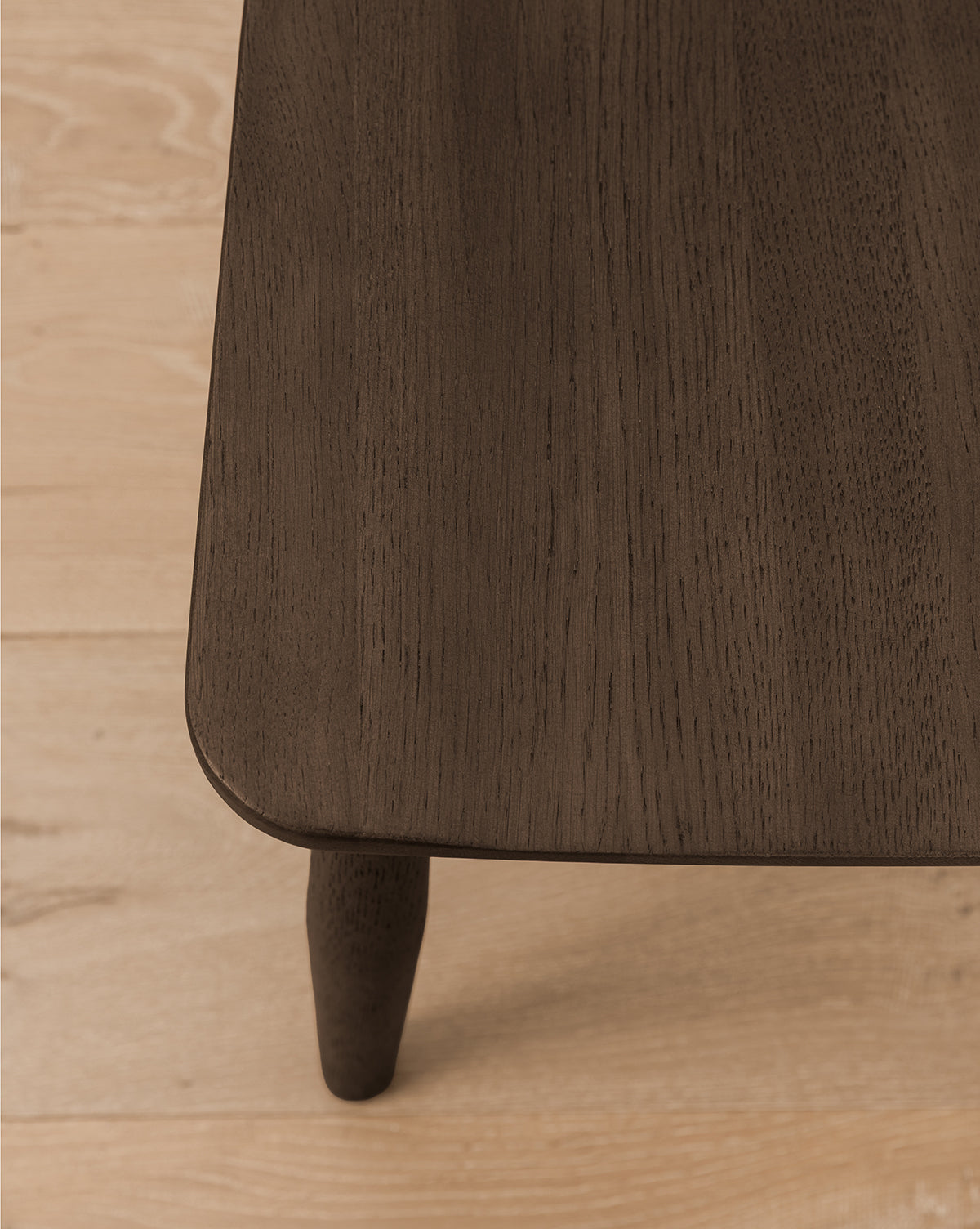 A close-up of the corner of a solid wood table with rounded edges and a visible leg on a light wood floor—an elegant accent beside kitchen counters or perfectly paired with the Hemingway Stool, by Vendixs
