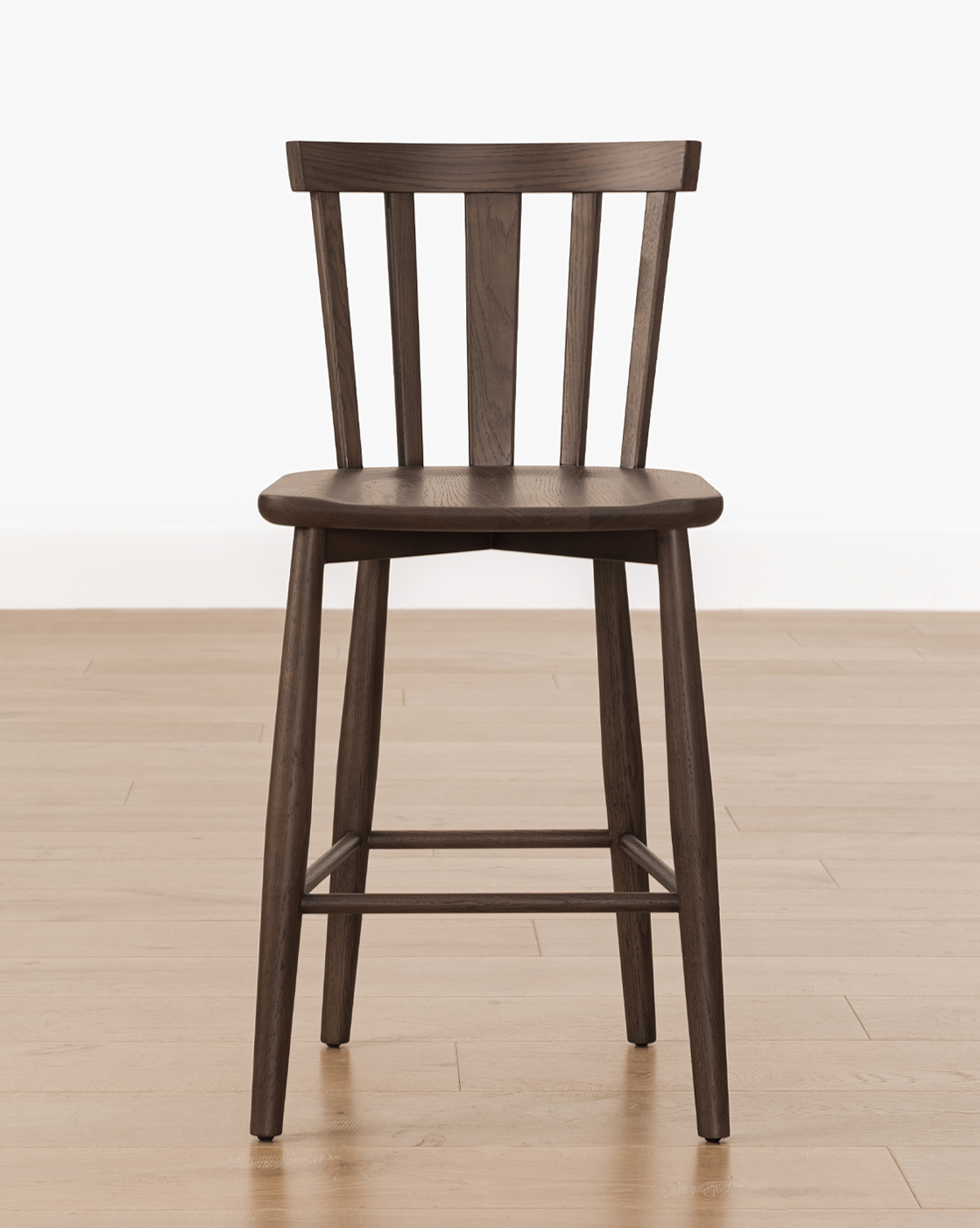 The Hemingway Stool is a dark solid wood chair with a simple design, featuring vertical slats on the backrest and four angled legs—perfect for kitchen counters, shown here on a light wooden floor against a plain white wall, by McGee & Co.
