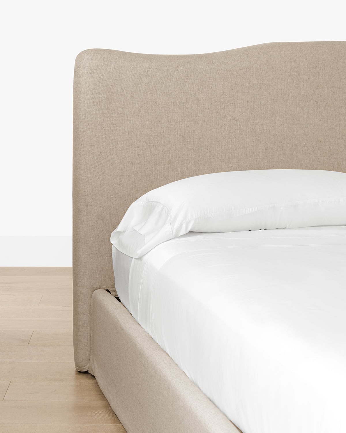 A close-up of the Henrietta Bed (Ready to Ship) features a beige upholstered frame with a tall, curved headboard, styled with white sheets and pillow on a light wood floor against a white wall for timeless bedroom appeal, by Vendixs