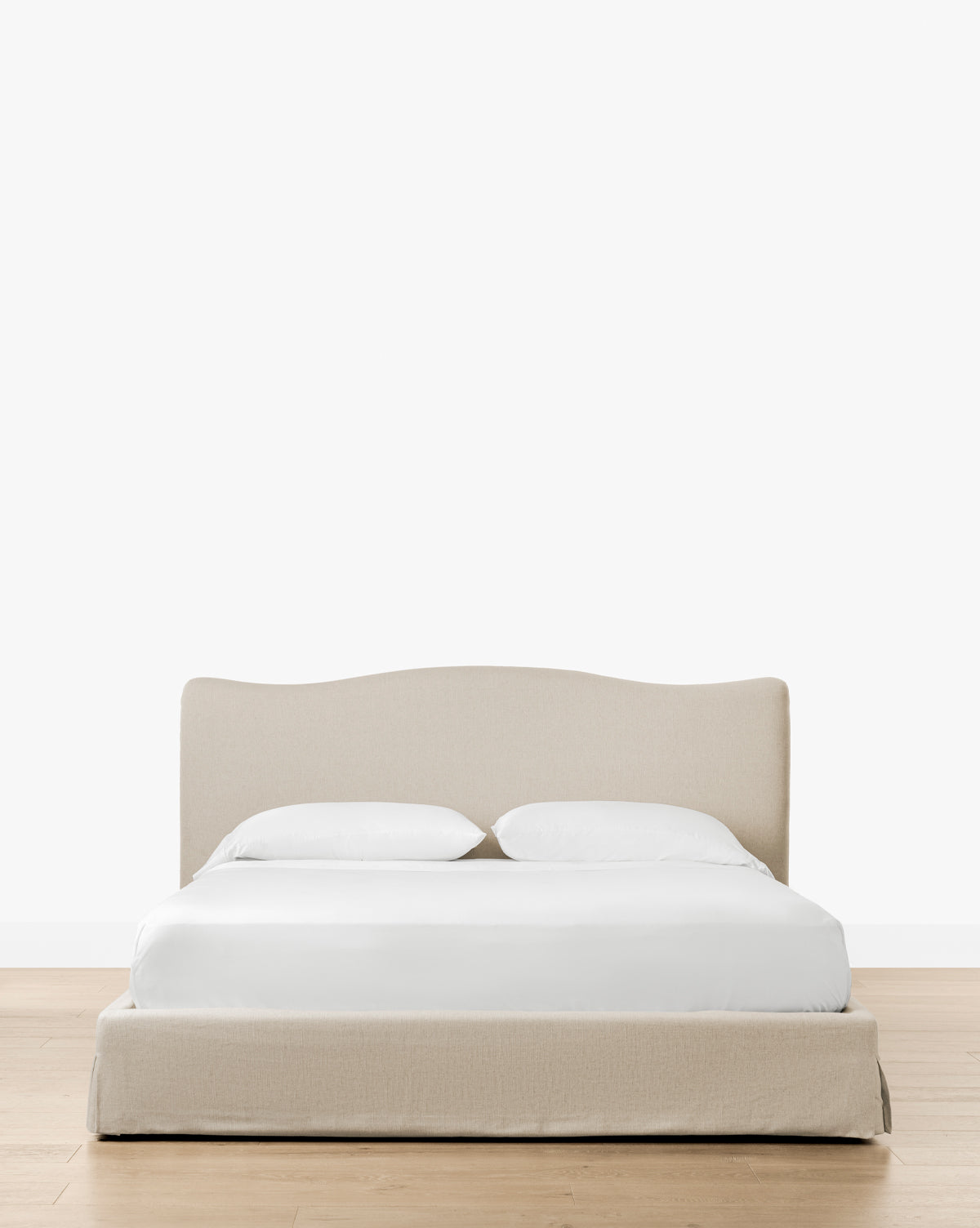The Henrietta Bed (Ready to Ship), featuring beige upholstery, a curved headboard, white bedding, and two pillows, sits on a light wood floor with a plain white backdrop—ideal for refined bedroom styling by Vendixs
