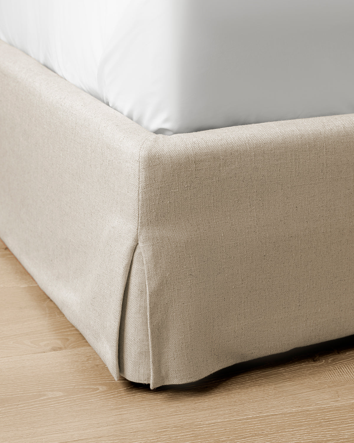 Close-up of the Henrietta Bed (Ready to Ship), showcasing its beige upholstered base with a neat corner pleat and a white mattress, set on a light wooden floor—an ideal choice for stylish bedroom updates, by Vendixs