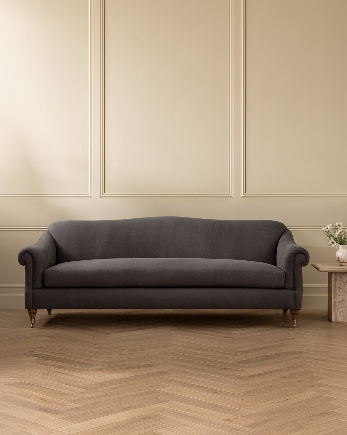 A dark gray Hildegard Sofa 90 with rolled arms sits against a beige paneled wall on a herringbone wood floor. To its right, a small stone side table holds a vase of flowers next to the handcrafted sofa, by Vendixs