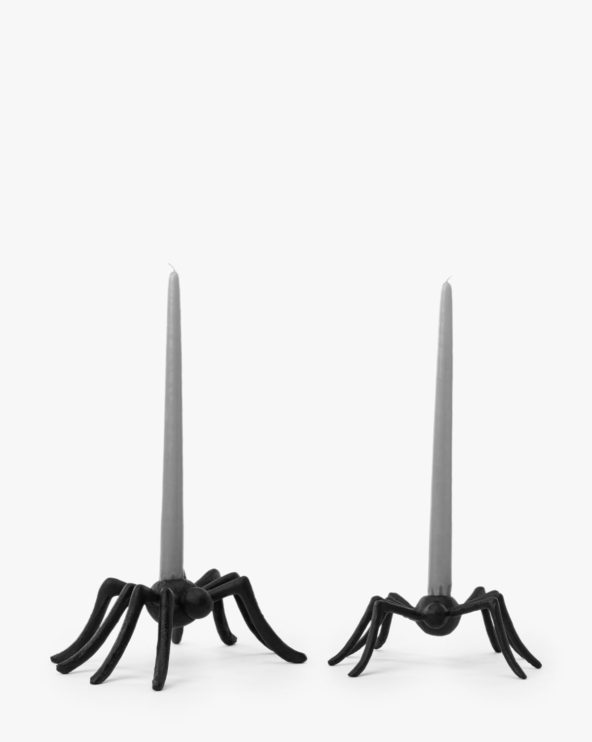 Two Iron Arachne Taper Holders, shaped like lifelike black spiders with spread legs, each hold a tall gray taper candle. These unique holders make striking Halloween decor against a plain white background, by Vendixs