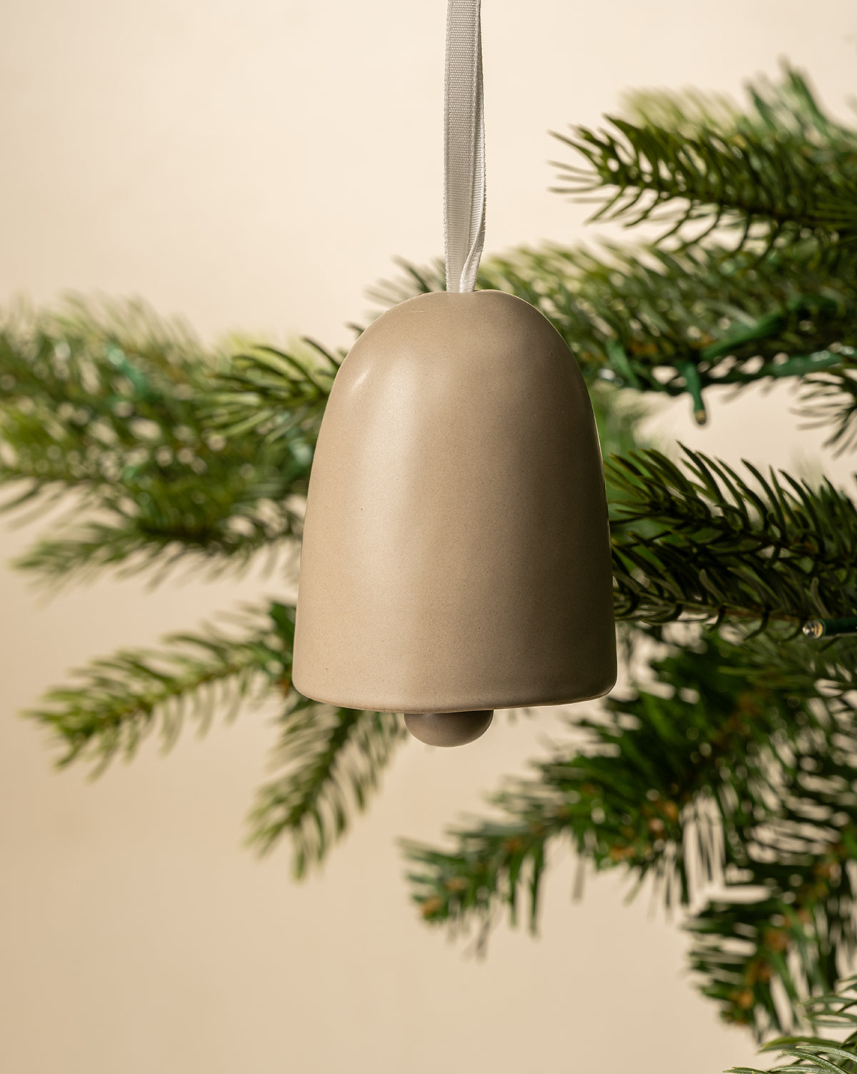 The Ivory Ceramic Bell ornament, hanging by a white ribbon on a green pine branch, adds classic holiday charm against a neutral backdrop, by Vendixs