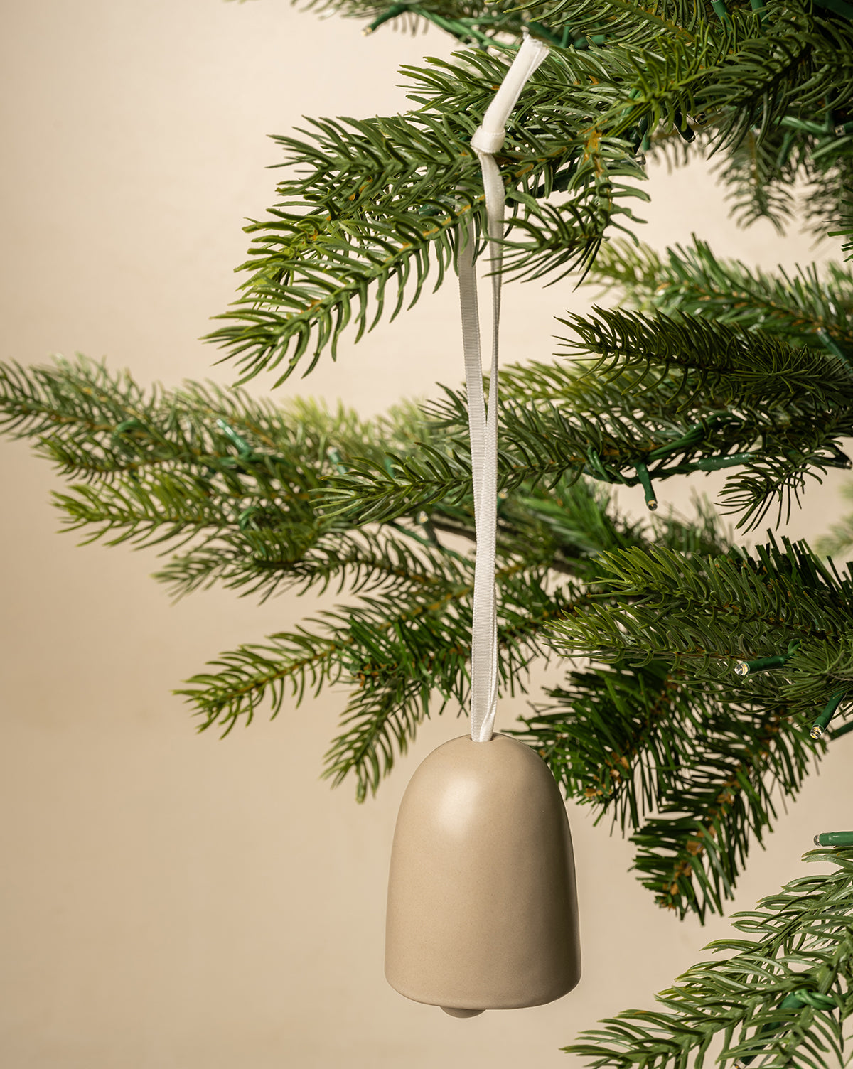The Ivory Ceramic Bell hangs from a white ribbon on a green pine branch, bringing classic charm to your holiday decor with its simple design and elegant ivory finish, by Vendixs