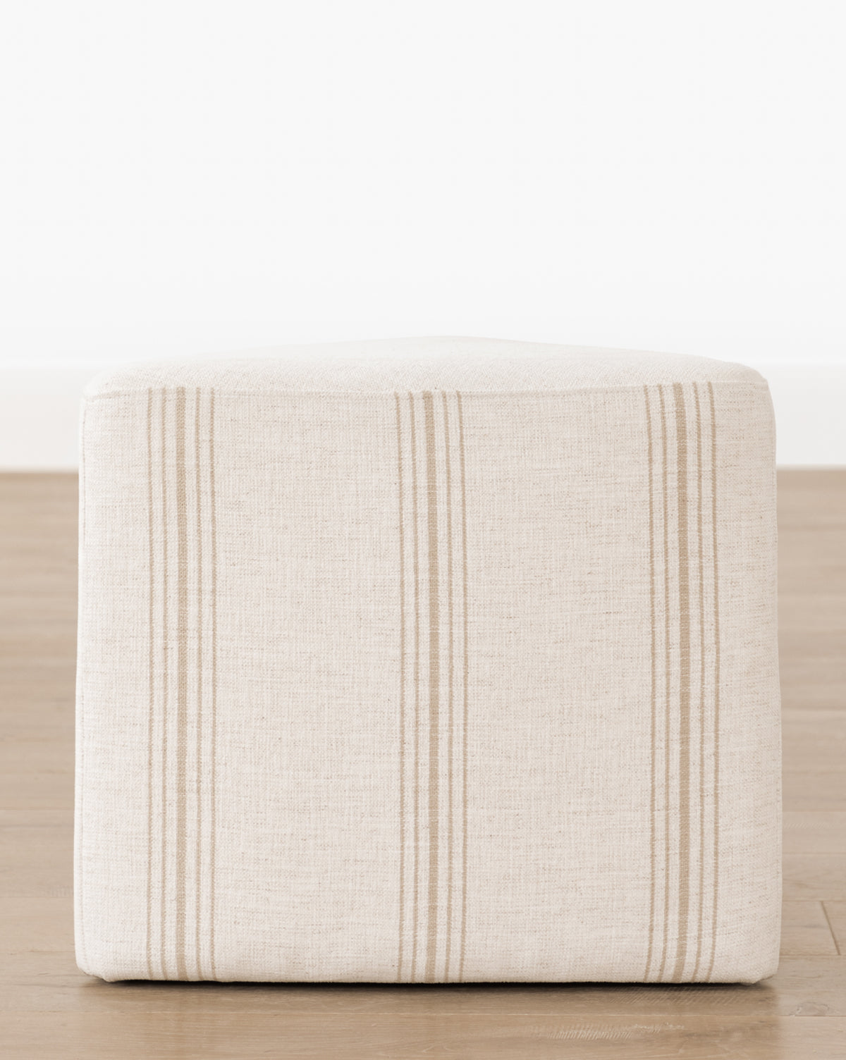 The Jacques Ottoman by Dongguan City Baby Furniture Co., Ltd features beige Crypton striped fabric in a cube shape with tan stripes—ideal for stylish, functional kids’ rooms. Shown on a wooden floor against a white wall. Vendixs