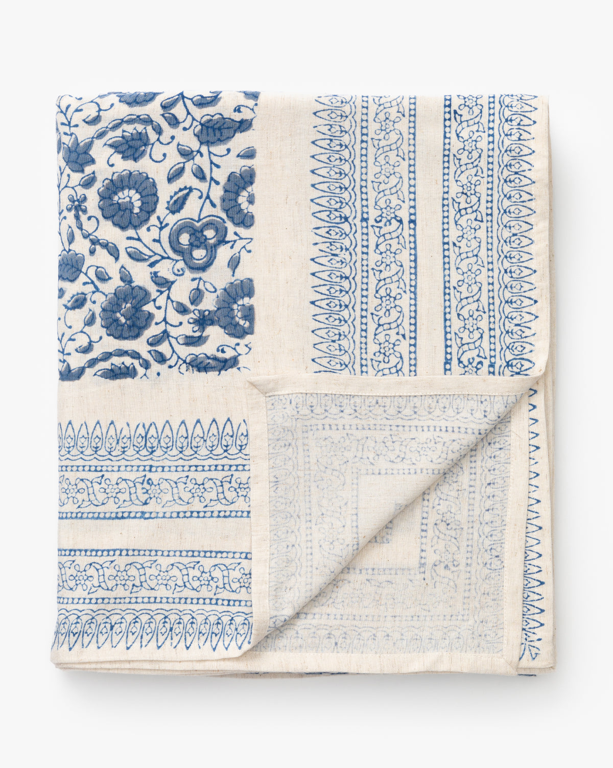 The Jennie Tablecloth is a folded cream-colored block printed tablecloth with a patchwork of intricate blue floral and geometric patterns, one corner neatly turned up to reveal the reverse side, by Vendixs