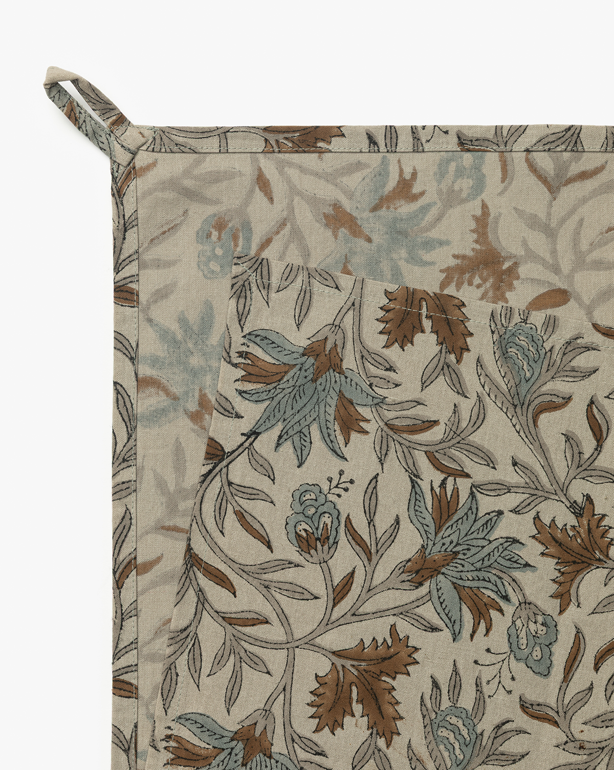 A close-up of a beige Jennifer Block Print Tea Towel (Set of 2), featuring blue-green and brown leaves and flowers in block print. Made of cotton with stitched edges and a fabric loop, it can be used as a napkin or placemat, by Vendixs