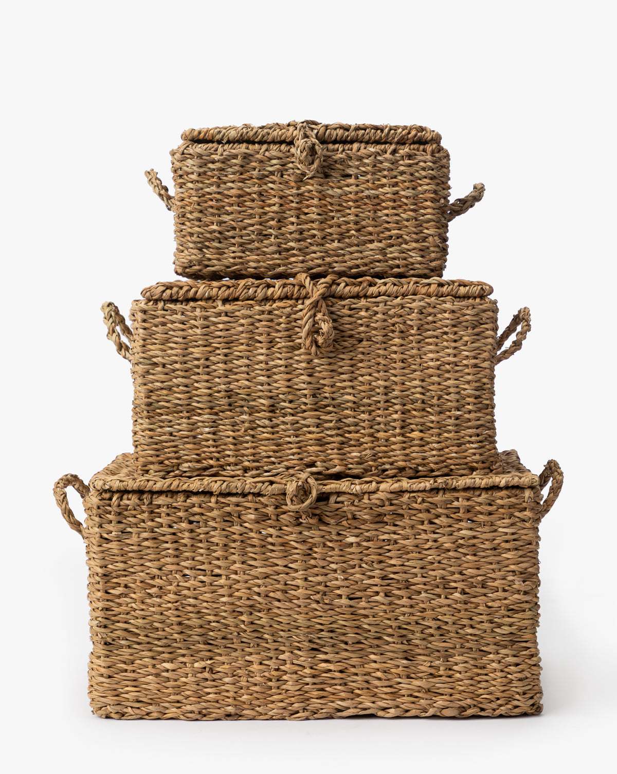The Kya Seagrass Box set features three rectangular, hand-woven wicker baskets with lids and side handles, stacked from largest to smallest for a charming decorative touch against a plain white background, by Vendixs