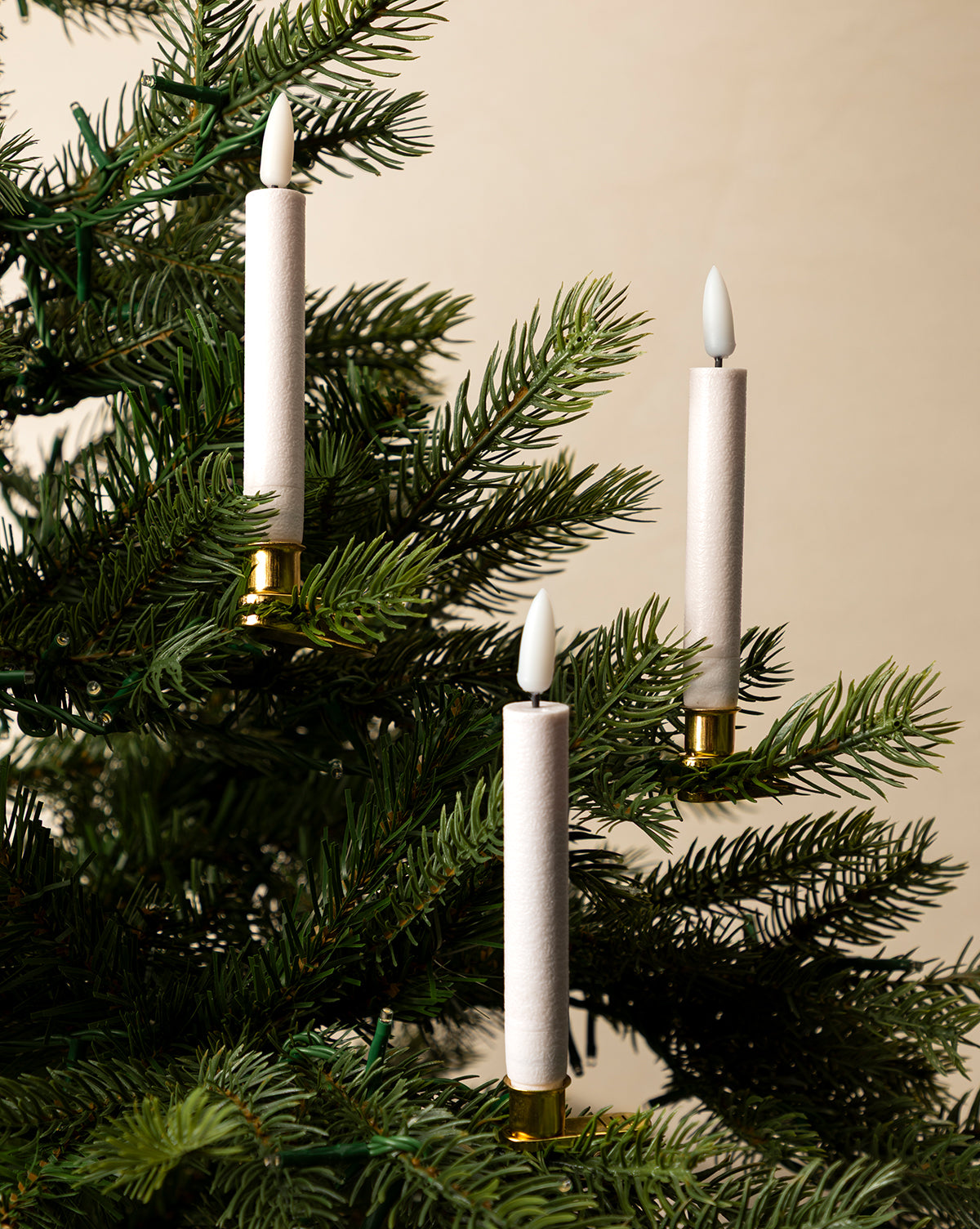 A set of 10 LED Candle Lights with Brass Clips are attached to the branches of a green Christmas tree, creating a festive holiday atmosphere against a soft beige background, by Vendixs