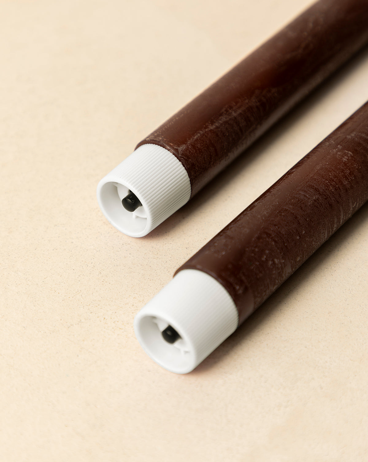 A set of two brown LED Wax Taper Candles with white caps and small black circular openings, lying parallel on a light beige surface, by Vendixs