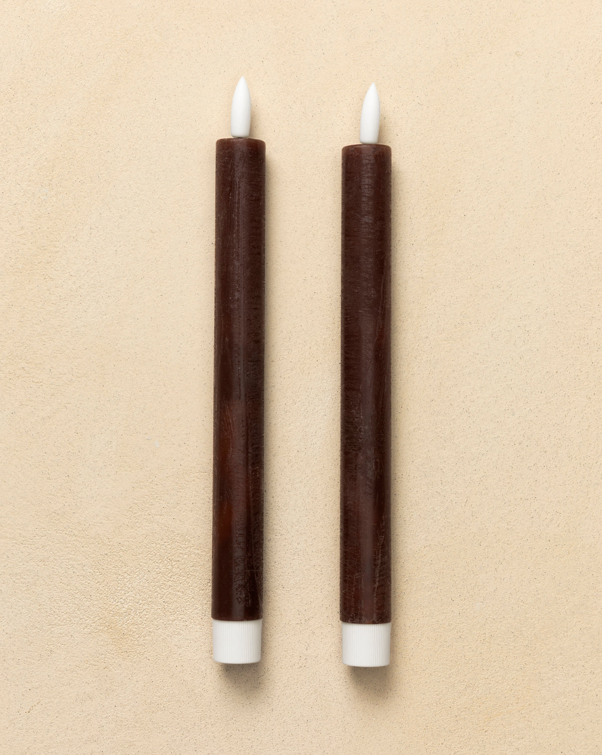 Two brown LED Wax Taper Candles (Set of 2) with white tips and bases stand upright on a light beige surface, providing the cozy glow of flameless candles, by Vendixs