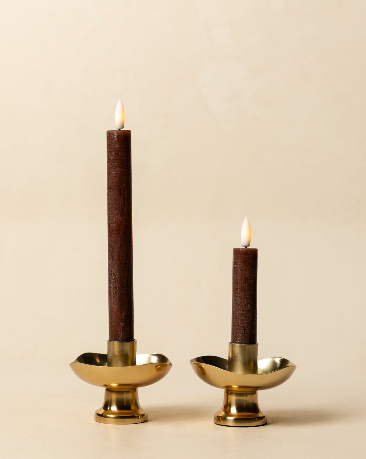 The LED Wax Taper Candles (Set of 2), in warm brown and varying heights, sit in gold holders on a beige background, offering the cozy glow of real tapers with safe, elegant LED flicker, by Vendixs