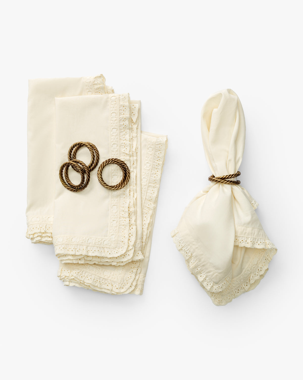 Four folded cream-colored Lace Trim Napkins (Set of 4) with delicate lace edges are stacked on the left, topped with three gold napkin rings. On the right, a single napkin is wrapped in a gold ring, all arranged on a white background, by Vendixs