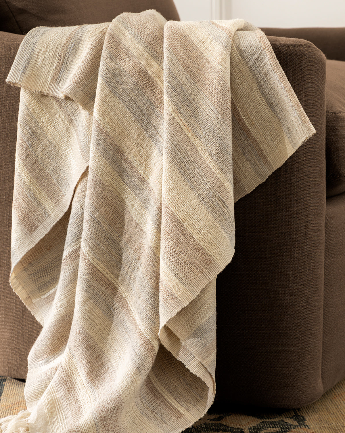 The Laney Stripe Throw by TAL HANDWOVEN CLASSICS LLP, in beige and cream stripes with tassels, is draped over a brown armchair, adding textured luxury and warmth to your space. Vendixs