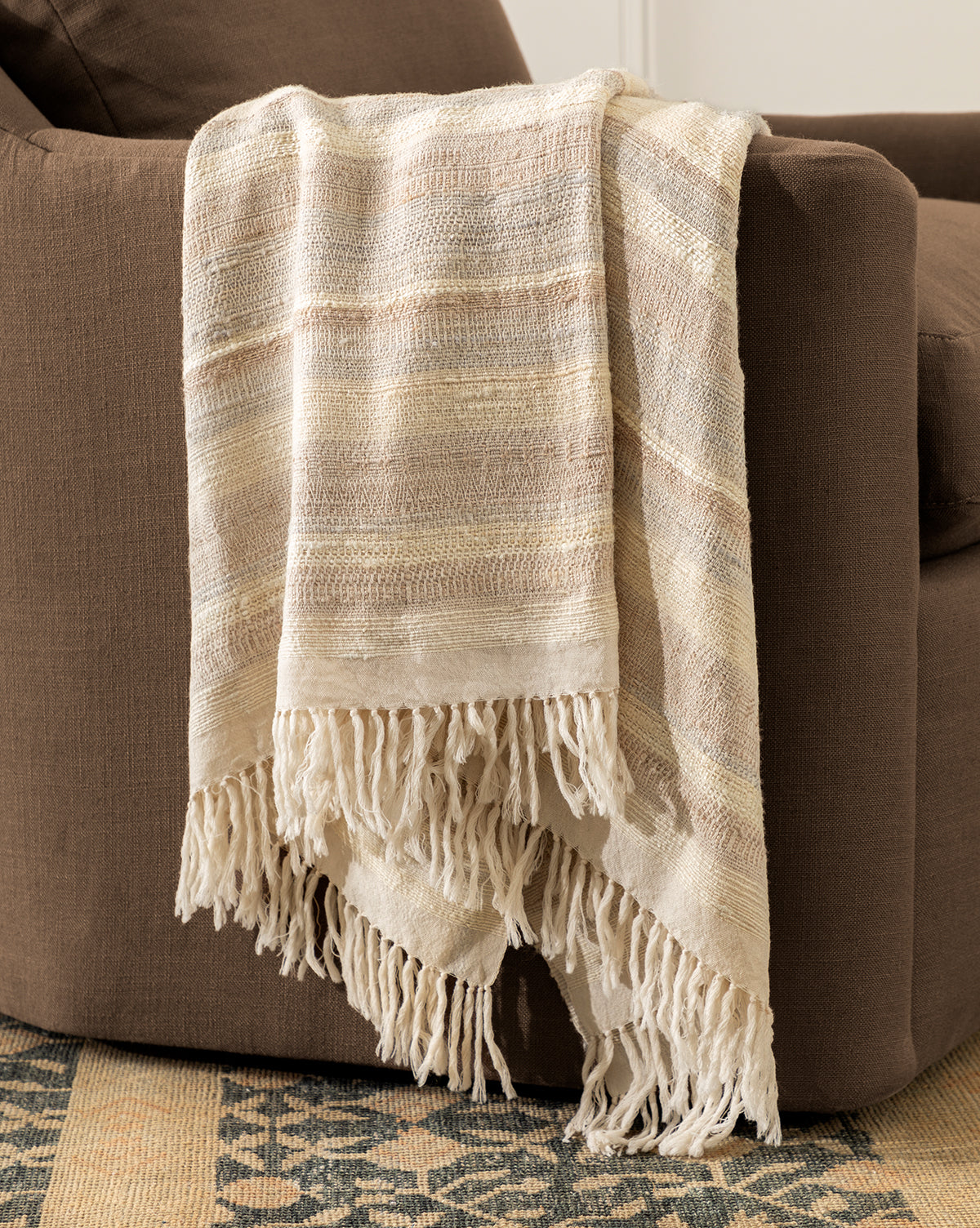 The Laney Stripe Throw by TAL HANDWOVEN CLASSICS LLP, featuring cream and beige stripes with fringed edges, is draped over a brown upholstered chair, with a patterned rug partly visible on the floor. Vendixs