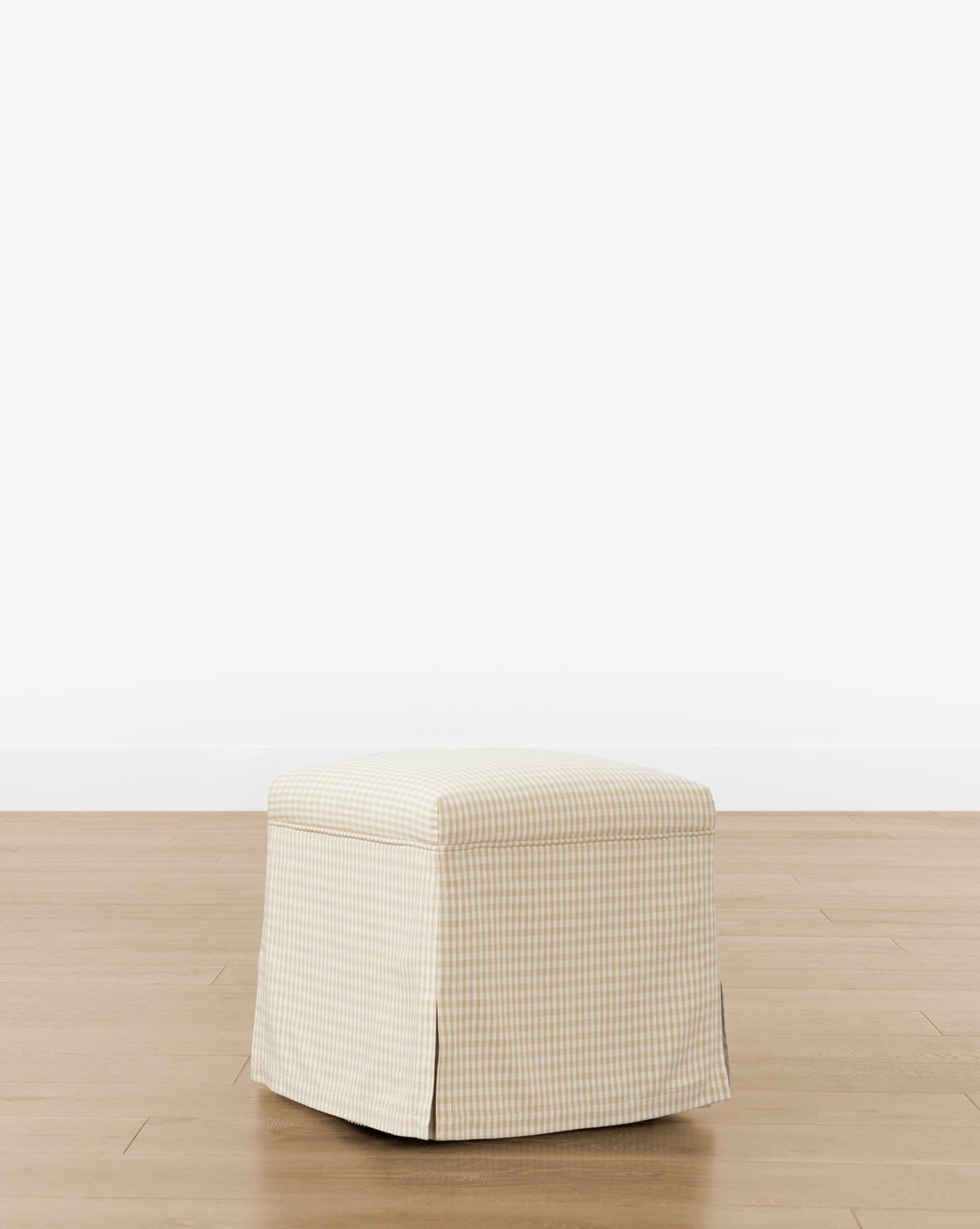 The Makerspalm Latimer Ottoman (Ready to Ship) features a beige and white gingham-patterned slipcover with pleated corners, shown on a light wood floor against a plain white background. Vendixs