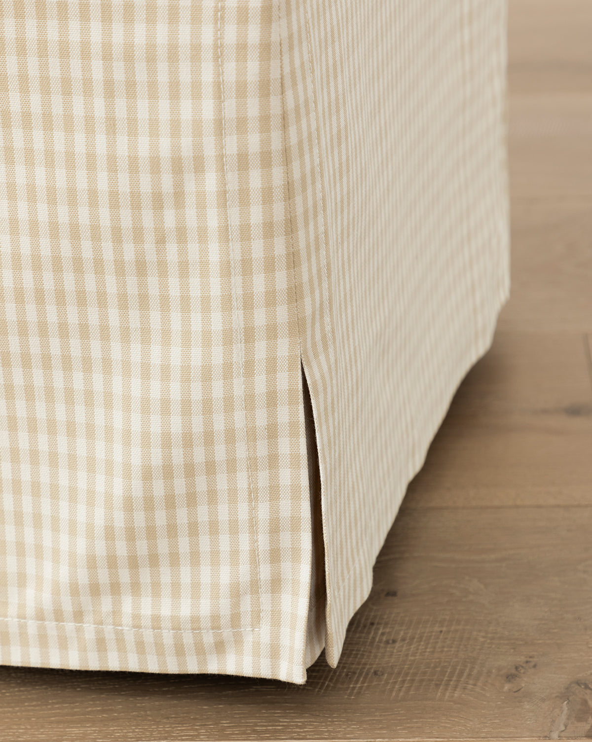 Close-up of the Makerspalm Latimer Ottoman (Ready to Ship), featuring beige and white gingham-patterned fabric with a tailored corner and vertical slit, elegantly placed on a light wooden floor. Vendixs