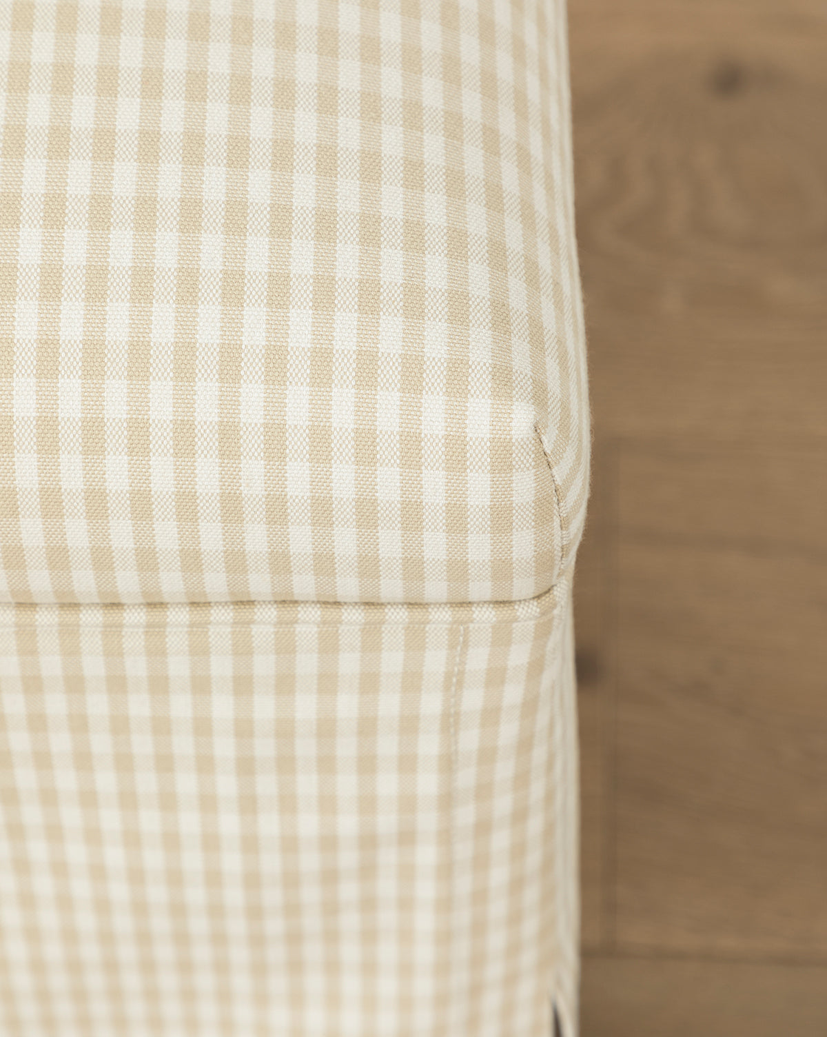 Close-up of the Makerspalm Latimer Ottoman (Ready to Ship), featuring beige and white checkered upholstery, showcases its quality craftsmanship as a stylish accent piece, set against a wooden floor. Vendixs