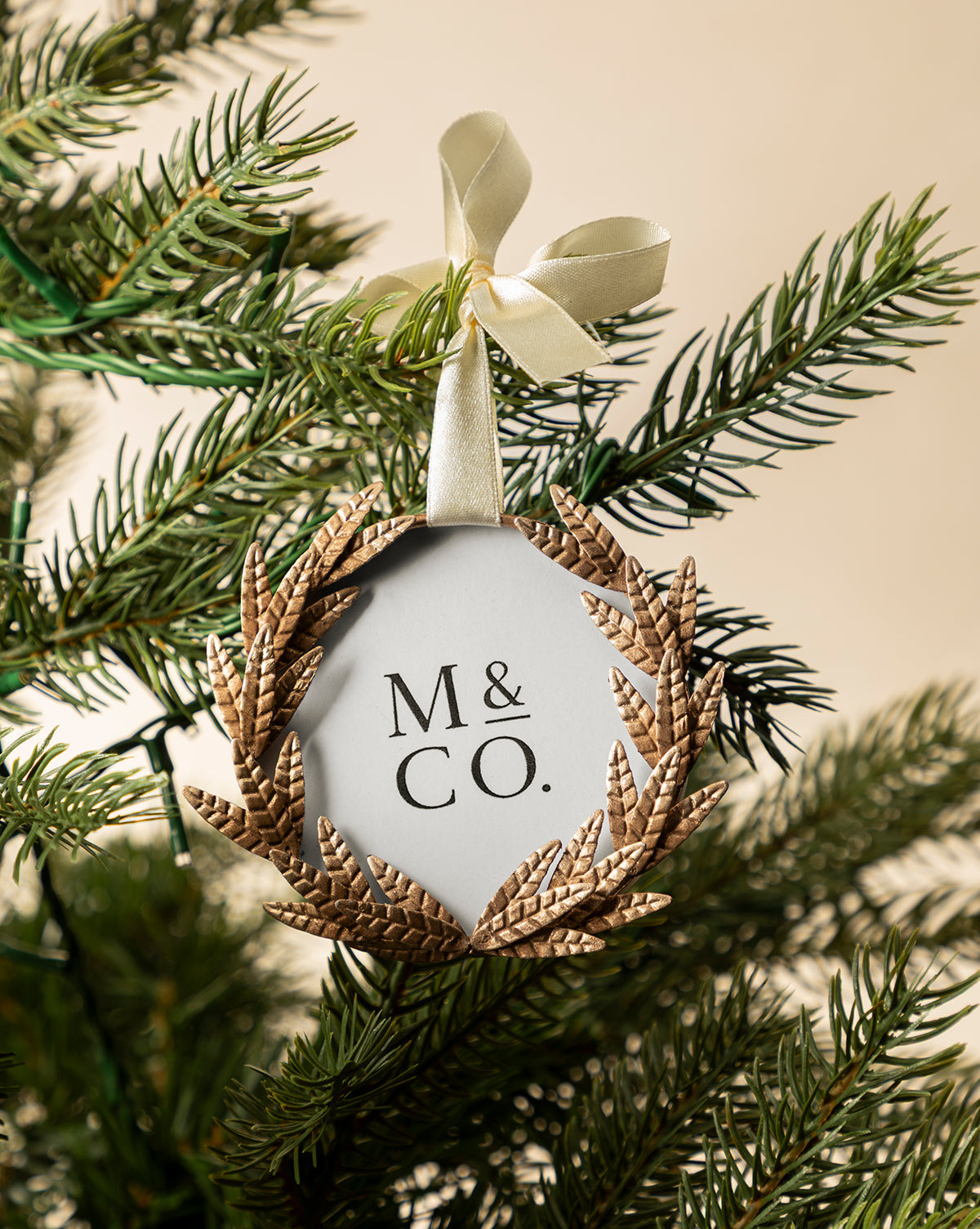 The Laurel Picture Frame Ornament features "M & CO." in black, framed by a gold laurel wreath and hung with a cream ribbon bow—an elegant, sentimental holiday gift that adds charm to any green Christmas tree branch, by Vendixs