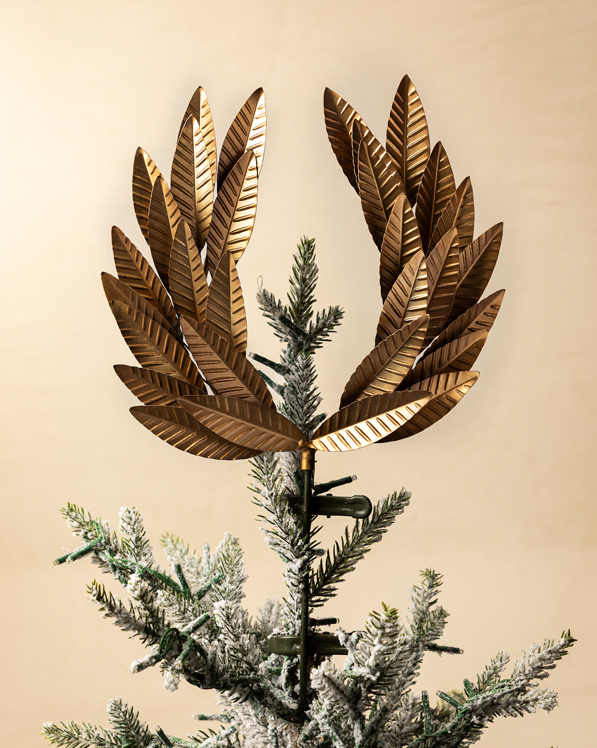 A close-up of a holiday tree features the Laurel Tree Topper in metallic gold, surrounded by green, snow-dusted branches set against a beige background, by Vendixs